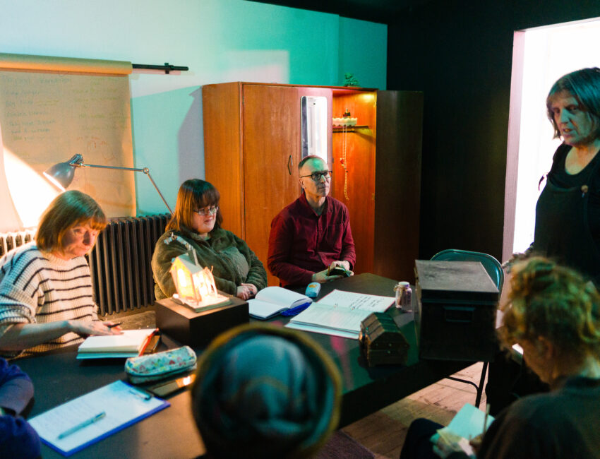 A group of adults writing. They are sitting around a table in a dark room