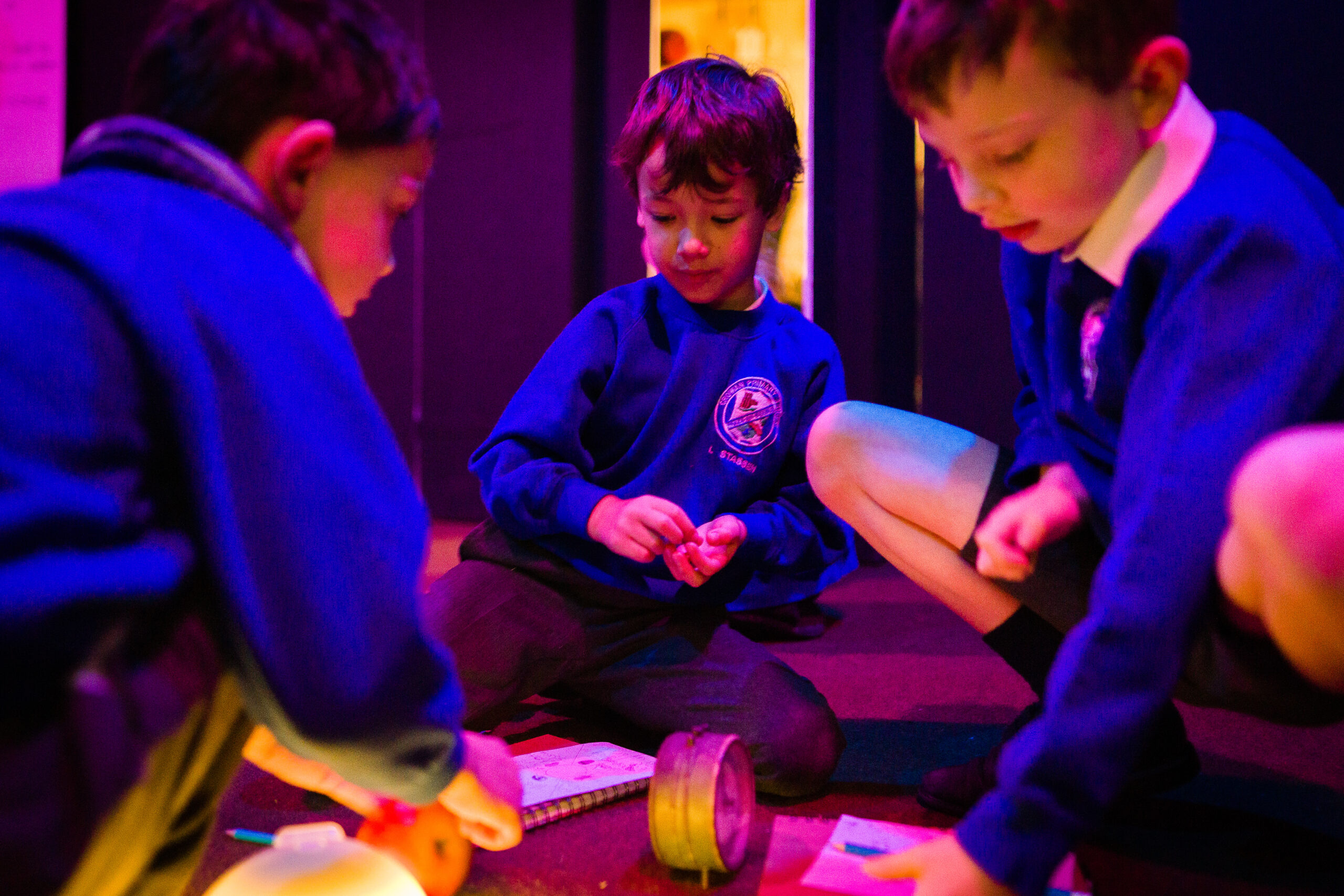 Three young children sit on the floor of a dark room looking at different objects and paper