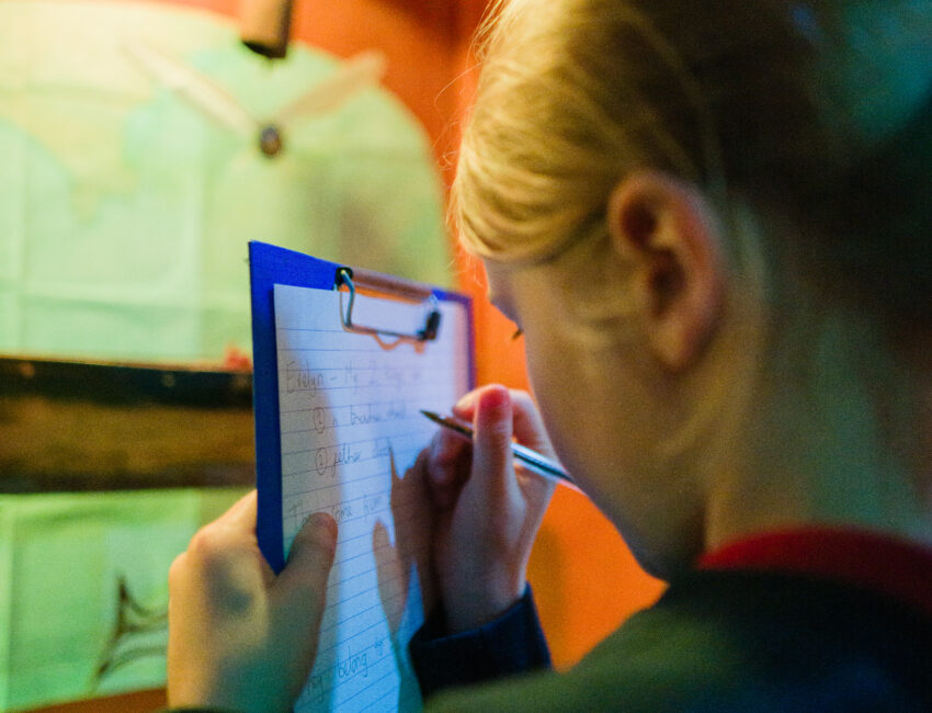 Child writes on a clipboard looking into a light up cupboard with props in it