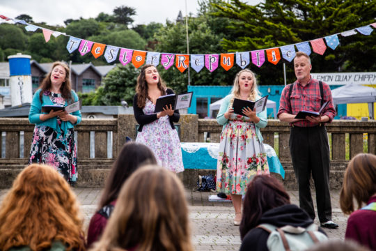 Four performers standing in a line with backs of audience heads in the foreground. Image by Sean Hurlock.